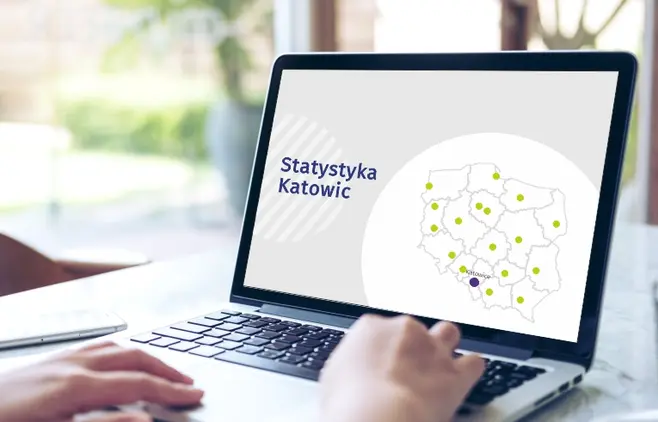 Otwarty laptop na biurku, na którego ekranie widnieje napis „Statystyka Katowic” oraz uproszczona mapa Polski z zaznaczonymi punktami wojewódzkimi, gdzie Katowice wyróżniono fioletową kropką.