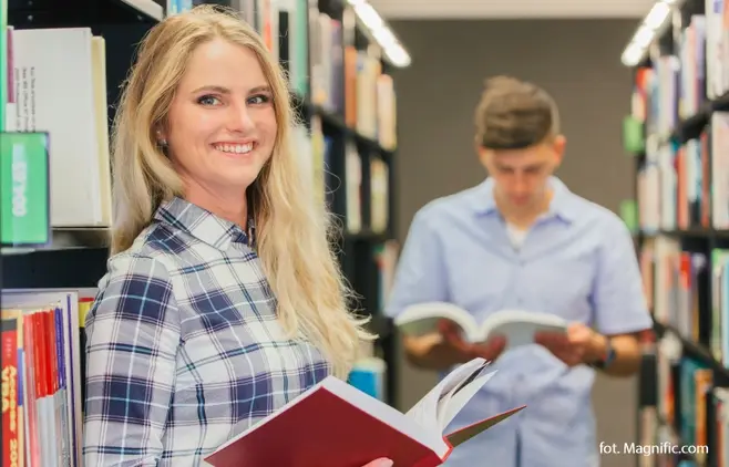 Dwoje uśmiechniętych, młodych ludzi w bibliotece uczelnianej, stoi w alejce pomiędzy regałami wypełnionymi książkami.